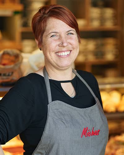 Micha, die Unternehmerin. Freundliches Lächeln, rotbraune, kurze Haare mit Sommersprossen. Schürze mit der Aufschrift Michas.